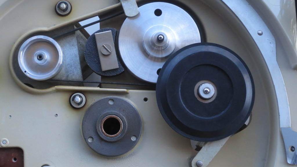 Underneath the platter(s), showing the spindle well (sintered bronze) at bottom centre. From upper left to right: reversible motor pulley (noting two very small balance holes), belt, stepped idler pulley and brand-new idler wheel. Note the large magnet next to the idler pulley; using the principle of eddy-current braking, fine control of speed is achieved.